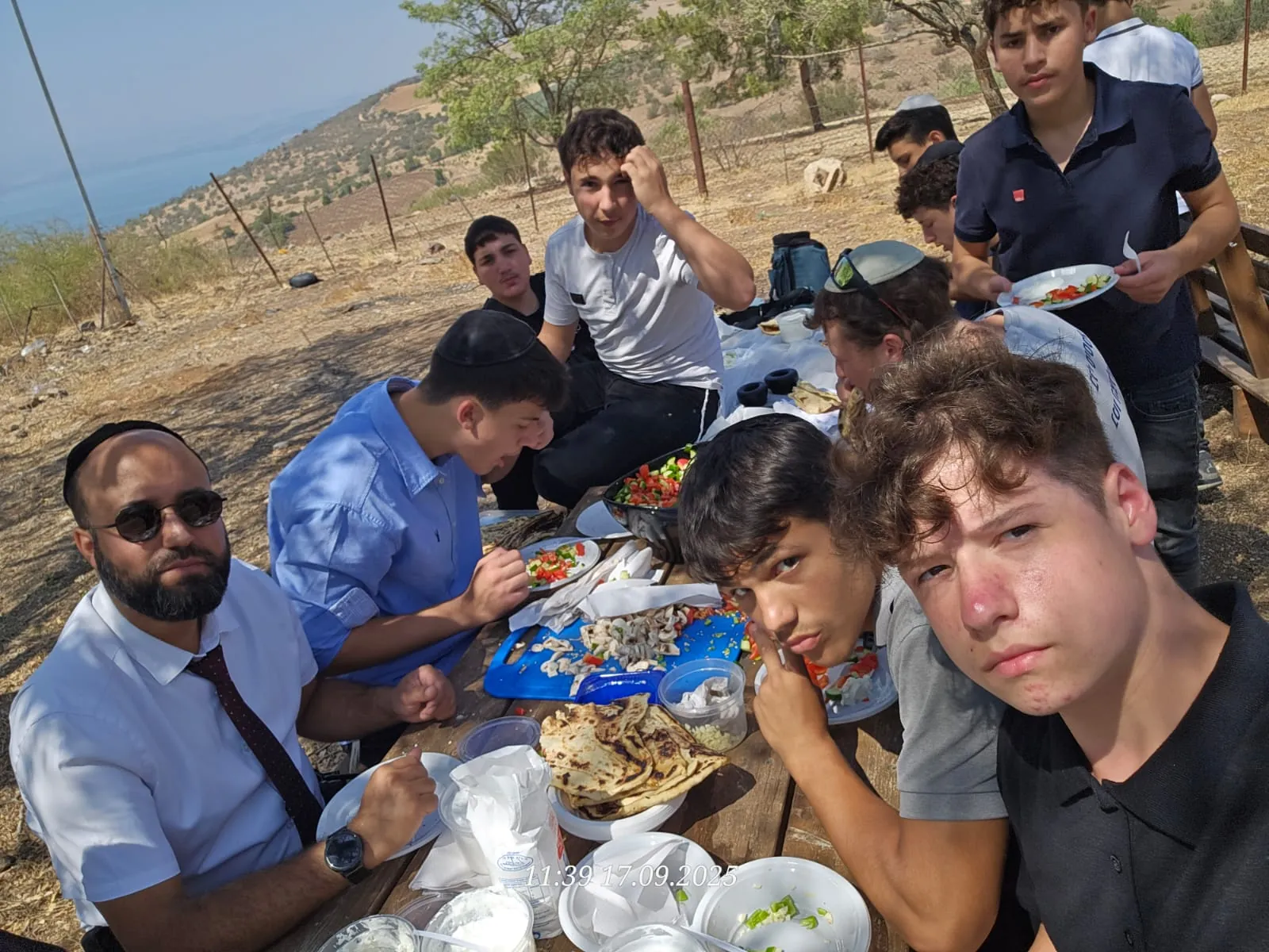 תלמידים ואיש צוות אוכלים יחד סביב שולחן בשטח - פיתות על המדורה - בוקר כיתה יא בחורשה