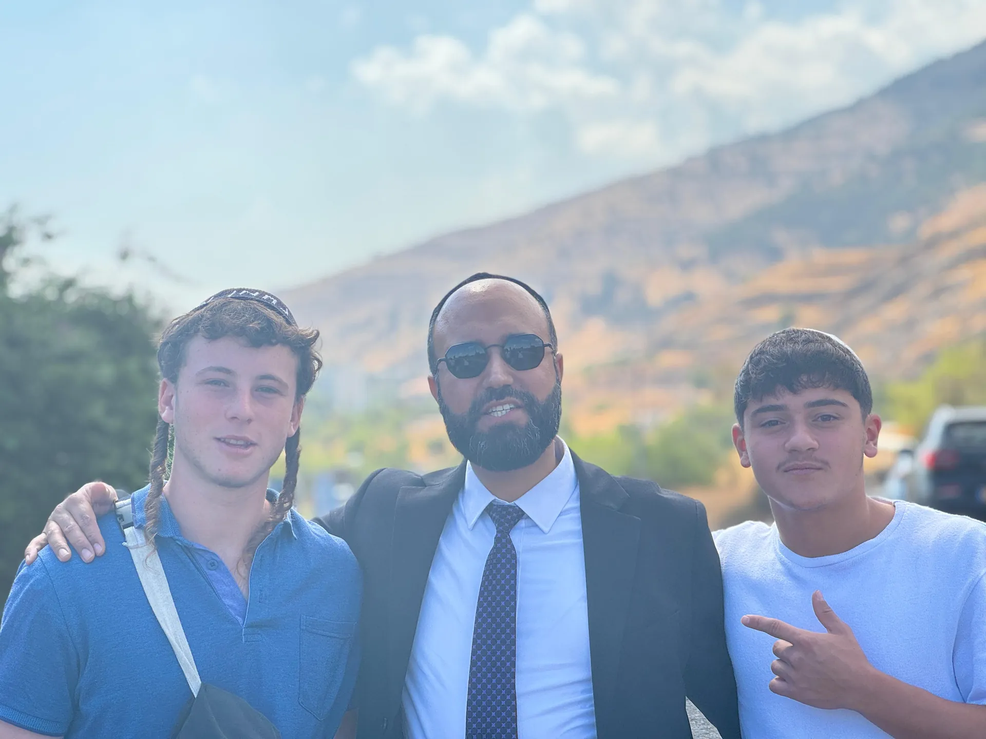 רב ושני תלמידים מצטלמים בחוץ מול נוף הרים - ערב ראש השנה בקברי צדיקים