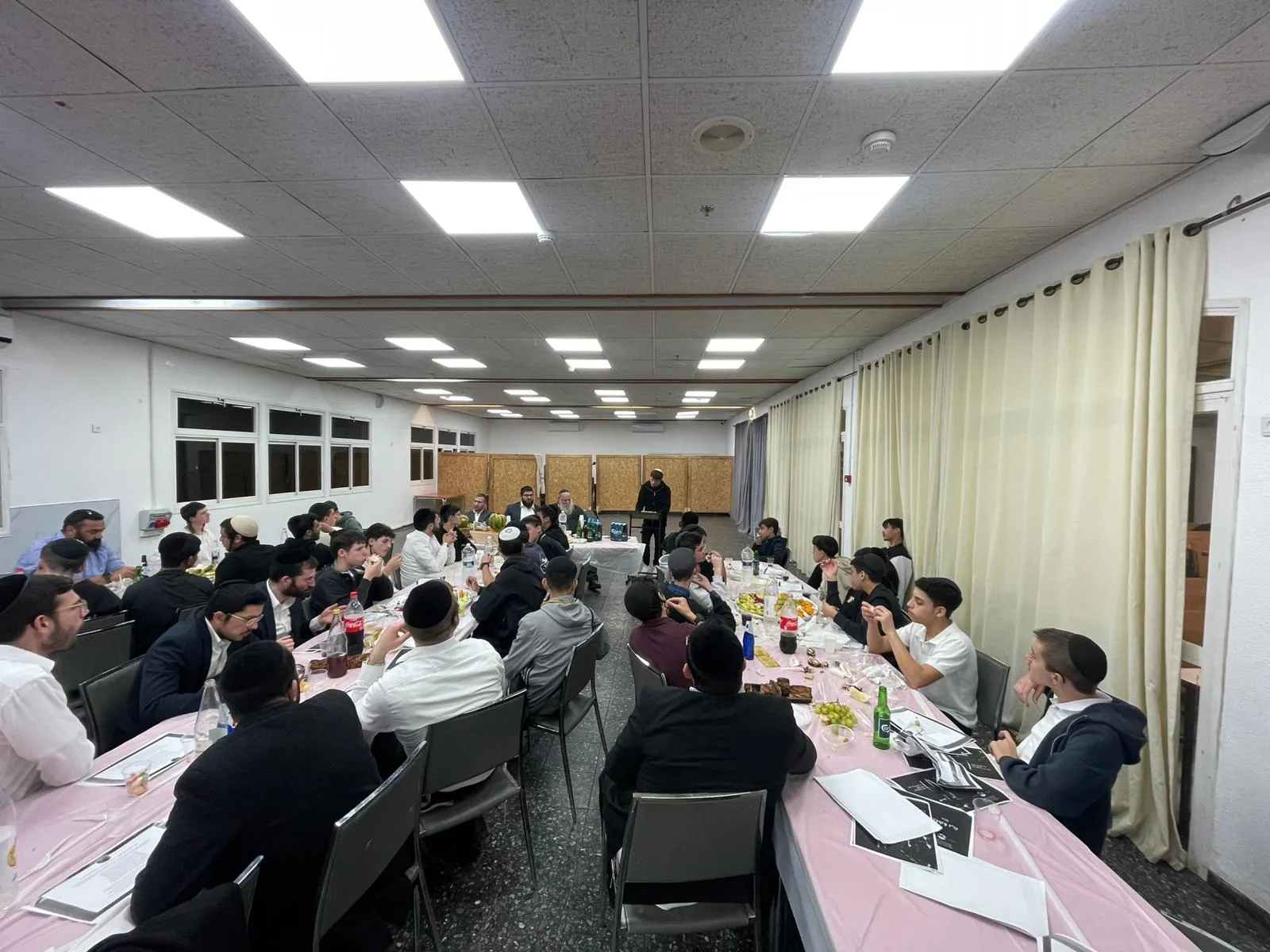 תלמידים ואנשי צוות יושבים סביב שולחנות סעודת טו בשבט - סעודת ט״ו בשבט