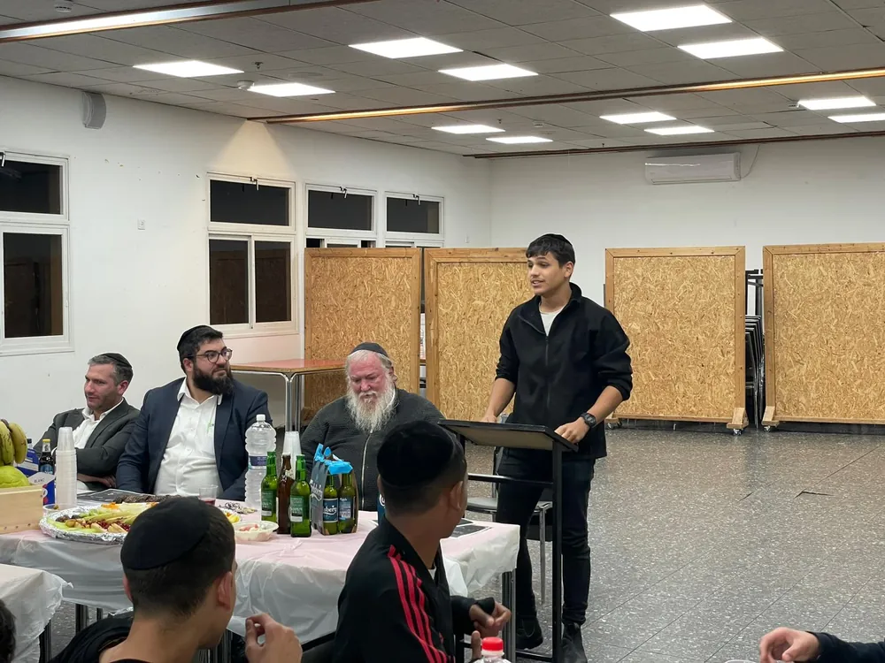 תלמיד בחולצה שחורה מדבר ליד שולחן טו בשבט - סעודת ט״ו בשבט