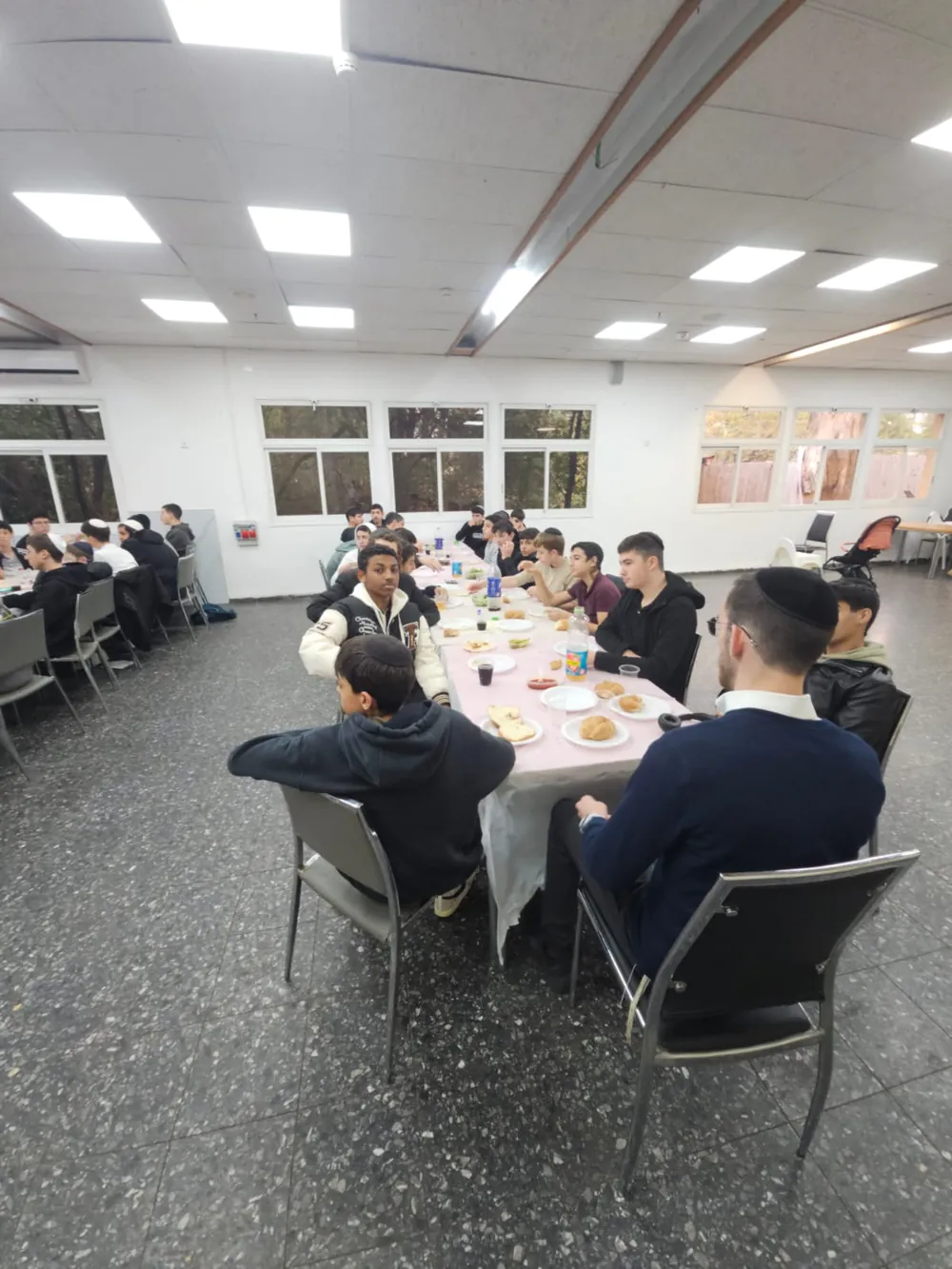 קבוצת תלמידים יושבת סביב שולחן סעודה עם לחמניות - סיום מסכת - תעודה ביד וסעודה משותפת